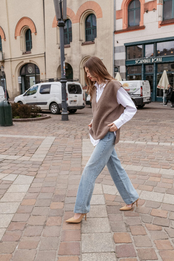 Žena v hnědé vlněné vestě, bílé košili a širokých světle modrých džínách kráčí po dlážděné ulici ve městě.