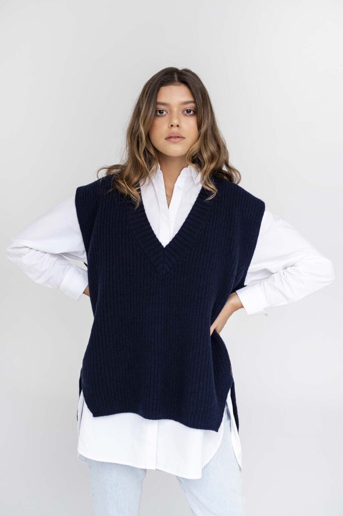 Žena v tmavě modré vlněné vestě, bílé košili a modrými kalhoty, s vlnitými vlasy.