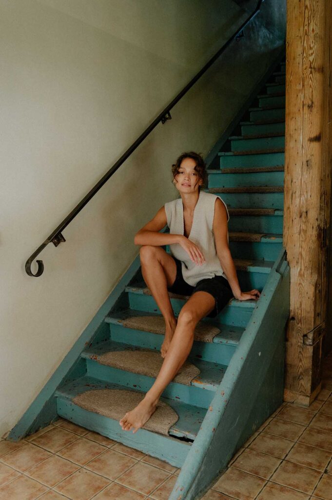 Žena v béžové vlněné vestě s tmavě modrými šortkami sedí na schodech.