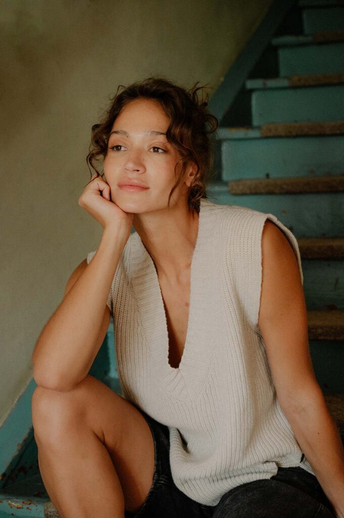 Žena v béžové vlněné vestě s tmavě modrými šortkami sedí na schodech.